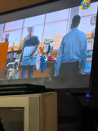 This is a medium-shot, eye-level, indoor scene, potentially a television broadcast, with a projected image of an office environment. The foreground features the edge of a television or projector screen with a black textured bar along the top and a beige-cased television set below. The projected image depicts two individuals in what appears to be an office or storage area. On the left, a person with short, gray hair, wearing a dark blue t-shirt and blue pants, is carrying a light-colored folder with papers inside and a pinkish-purple bag. To their right, a young child in a light-colored outfit is partially visible. On the right side of the projected image, a person wearing a light blue collared shirt and dark pants stands with their back to the camera, facing away. Behind them, shelves are filled with binders, boxes, and what appears to be office equipment, including a printer. The background features large windows with blinds. In the upper right corner of the projected image, a colorful, cube-shaped graphic with the letters "TBB HD" is visible. The overall lighting appears to be artificial, typical of an indoor setting. There is no identifiable information in the image to suggest the location is the city of Chorbogh, Tajikistan.