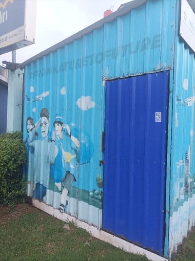 The image displays a light blue, corrugated metal structure, resembling a modified shipping container, positioned on a grassy patch under an overcast sky. A prominent mural covers the structure, featuring the faded black text "#FROMNATURETOFUTURE" across its upper section. Below the text, stylized cartoon figures are depicted: a female on the left, a male with glasses in the center, and another male on the right, all appearing in motion. The background of the mural includes white clouds, a faint depiction of a Garuda Indonesia airplane, and a green landscape at the bottom. A royal blue, corrugated metal door is centrally located on the structure's front face, secured by a dark handle and a red-tipped latch. A small white sticker containing a QR code is affixed to the door. To the immediate left of the structure, dense green shrubbery is visible. In the upper left corner, a white vertical sign, partially obscured, displays the "mandiri" logo, "e-money," and the term "blu," indicating a connection to Indonesian financial institutions like Bank Mandiri and Bank BCA Digital. Another partial white sign is visible on the upper right side of the structure. The ground in the foreground is covered by green grass. The structure exhibits some weathering, with minor paint scuffs and dirt accumulation, particularly at its base. The presence of specific Indonesian banking and airline branding strongly indicates the location is within the Special Capital Region of Jakarta, Indonesia, likely in a commercial or public access area.