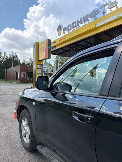 A black SUV is parked at a gas station with a large yellow sign reading "РОСНЕФТЬ" (ROSNEFT) overhead. The gas station appears to be in a rural or suburban area, as trees are visible in the background. The sky is partly cloudy. There are no people visible in the image. The focus is on the car and the gas station.