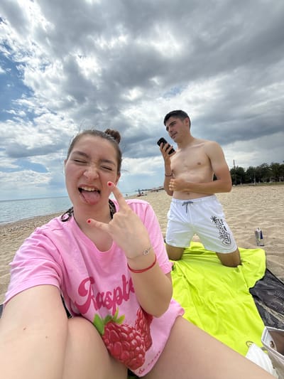 On the sandy beach of Νέα Φλογητά, Greece, two individuals pose for a photo. A woman in a pink "Raspberry" T-shirt is in the foreground, sticking her tongue out and making a hand gesture. Behind her stands a shirtless man in white shorts with "California" printed on them, looking at a smartphone. They are on a yellow beach towel, and the sky is overcast with blue patches.
