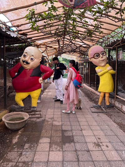 The image captures a scene at the "Motu Patlu" themed park in Nashik, India. It shows life-sized statues of the characters Motu and Patlu flanking a walkway with pedestrians.

Motu, in a red shirt, yellow pants, and black shoes, is positioned on the lower-left side of the frame. Patlu, wearing a yellow kurta and black shoes, stands on the upper-right.

Between the statues, several individuals are walking along a tiled path. One person wears a pink salwar kameez and carries a red backpack. Two other individuals are seen wearing white pants and black jackets. Overhead, a canopy of branches and a woven structure provide shade. A decorative umbrella is visible on the left. The surrounding environment consists of paved walkways, foliage, and constructed structures.
