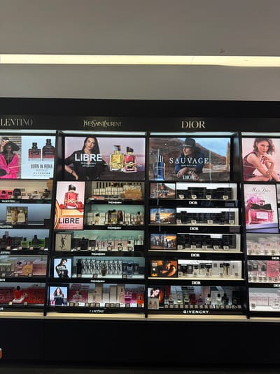 The image shows a retail display of perfumes and colognes in what appears to be a department store or duty-free shop. The display is organized by brand, with large illuminated signs for "VALENTINO", "YVES SAINT LAURENT", and "DIOR" at the top. Below these brand signs, numerous shelves are filled with various perfume bottles, boxes, and promotional images of models and product shots.
Specifically, the left side of the display features "VALENTINO" products, including bottles and imagery related to "BORN IN ROMA". The central section showcases "YVES SAINT LAURENT" fragrances, such as "LIBRE", with promotional posters of models. The right side is dedicated to "DIOR", with prominent displays for "SAUVAGE" and "Miss Dior". Further down, "GIVENCHY" and "LANCOME" are also visible.
The lighting is bright, illuminating the products and the brand signage. The environment appears to be an indoor retail space with a clean and organized presentation of merchandise. The image does not provide any specific indicators of its location within Xuanwu District, China. There are no individuals visible in the image.