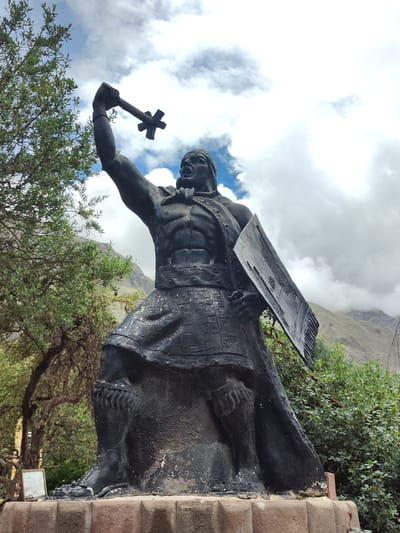 A dark stone statue stands heroically, holding a weapon overhead and a shield to its side. The figure is muscular and clad in traditional attire. A backdrop of mountains partially obscured by clouds adds depth, while a leafy tree and shrubbery surround the monument. The statue rests on a stone pedestal, blending art and nature in a scenic location.