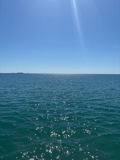 The image shows a view of the Mediterranean Sea from Fréjus, France, on a clear, sunny day. The sky is a gradient of blue, ranging from a lighter hue near the horizon to a deeper blue overhead. The sea is a textured expanse of water, with waves reflecting sunlight. A faint line suggests the presence of ships in the distance. The image is dominated by the expanse of sea and sky.