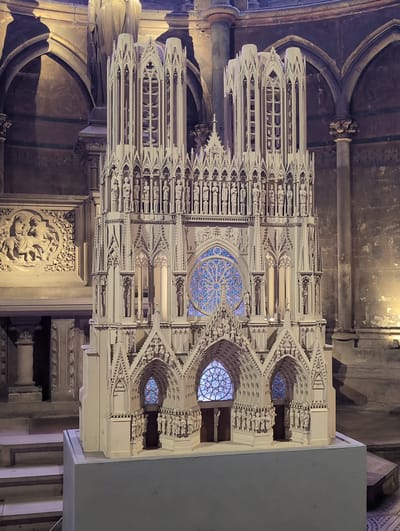 The image shows a detailed architectural model of a Gothic cathedral, likely a miniature replica. The model is constructed from a light-colored material, possibly plaster or resin, and features intricate carvings, statues, and stained-glass-like windows in shades of blue and purple. The model is positioned on a grey, rectangular base.
In the background, the interior of a larger, older building is visible, characterized by arched stone walls, columns, and decorative carvings. A large, relief sculpture depicting figures on horseback is mounted on the wall to the left of the model. The lighting in the scene is dim, with spotlights illuminating the model and parts of the background architecture, casting shadows.
The question states that this image is from Urvillers, France. However, a visual analysis of the architectural style of the model does not directly confirm this location. The Gothic style of cathedrals is common throughout France and Europe. Without additional information or specific architectural markers that definitively link this model to Urvillers, it's not possible to confirm the location based solely on the image content. However, if we assume the provided location is correct, the model is likely on display within a building in Urvillers. There are no people visible in the image. The scene appears to be an exhibit or a display within a museum or historical site.