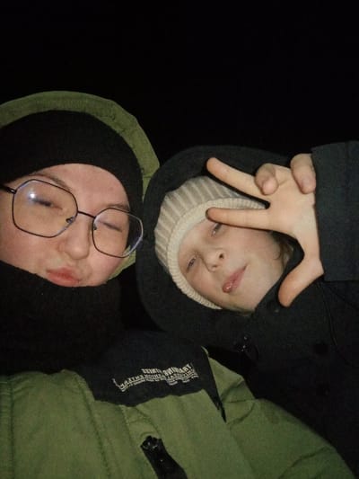 The image displays two individuals, an older youth/young adult and a child, positioned in a close-up, possibly selfie-style shot against a completely dark background, indicative of nighttime or extremely low light.
The individual on the left, an older youth/young adult, wears black-rimmed glasses, a black beanie, a black neck gaiter, and an olive-green hooded winter jacket with a black upper section bearing mirrored white text, including "TRAINING FITNESS." This person has pursed lips and faces the camera.
The individual on the right, a child, wears a light beige ribbed beanie beneath a dark hooded jacket. This person extends their tongue slightly and makes a "peace sign" (V-sign) with their right hand, partially obscuring their face, while also looking towards the camera.
Both individuals are dressed in warm clothing suitable for cold weather. The scene provides no discernible environmental context beyond darkness, and no specific features of the city of Старый Шагирт, Russia, are identifiable from the image content.