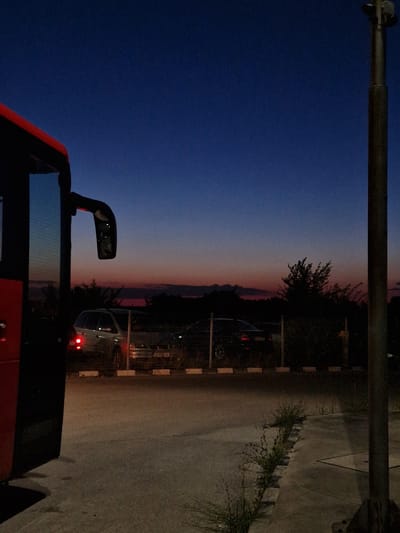 The image captures a roadside scene at dusk, with a large red bus occupying the left foreground. The bus's side is dark, with its reflection visible in the large side mirror mounted on the right side of its front. Behind the bus, a chain-link fence encloses a parking area. Two cars are parked within this area, their headlights and taillights illuminated. The car on the left appears to be a silver SUV, while the car on the right is darker. The ground is paved, with a curb separating the parking area from a slightly elevated sidewalk or walkway. Overgrown grass and weeds are visible along the edge of this walkway. In the background, a line of trees and foliage is silhouetted against the twilight sky, which transitions from a deep blue at the top to hues of pink and orange near the horizon. A tall, dark pole, possibly a lamppost, stands vertically on the right side of the frame. The overall atmosphere is quiet and transitional, characteristic of the hour between daylight and night. The location is identified as being in the city of Bahovitsa, Bulgaria.