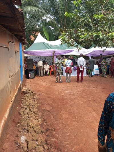 This image captures an outdoor gathering in Oko Town, Nigeria. The scene features a group of approximately 20-30 individuals assembled on reddish-brown soil under a large canopy tent with white and dark green fabric, accented by lavender drapes. Lush green tropical foliage, including palm trees, forms the background.
To the left, a single-story building with a corrugated metal roof presents a light brown, possibly rendered, wall with a vertical blue stripe and horizontal seams. A pile of loose, earthy debris is situated at the base of this wall. Two dark-colored loudspeakers, one atop another, are positioned near the building and the tent, with a black cable extending across the ground from their vicinity.
Under the tent, several people are seated on plastic chairs of various colors (red, orange, white). A white-covered table with purple floral arrangements is centrally located within the tented area. Various individuals are standing in the foreground and mid-ground: one man in a tan suit is bending forward, another in camouflage-patterned attire stands facing the tent, and a man in a white shirt and maroon trousers stands with his back to the viewer, holding a white paper. A woman in a vibrant patterned dress is partially visible on the far right. Attendees are dressed in a mix of traditional patterned clothing and Western semi-formal attire, engaged in social interaction typical of a community event.