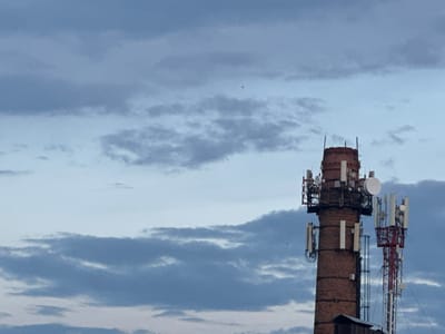The image shows two cell towers against a cloudy sky. The tower on the left is a brick structure with multiple antennas and dishes attached. The tower on the right is a red and white lattice tower, also equipped with various antennas. The sky is filled with a mixture of gray and light blue clouds, suggesting it might be overcast or nearing dusk. The lower portion of the image shows a hint of a dark structure at the base of the brick tower, possibly a building or shelter. There are no people visible in the image. The scene appears to be an industrial or urban area where communication infrastructure is located.