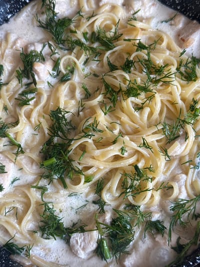 This media file is a close-up, overhead shot of a creamy pasta dish with chicken and dill. The pasta appears to be fettuccine or a similar flat noodle, coated in a rich, white sauce. Chunks of white chicken are interspersed throughout the pasta. Fresh green dill is generously sprinkled over the top, providing a vibrant contrast to the creamy tones of the dish. The setting appears to be a cooking pan, possibly on a stovetop, given the dark, textured edge visible on the left and right sides of the frame. The lighting is bright, suggesting daylight or good indoor lighting, and there are no discernible weather cues. The image focuses entirely on the food, giving no indication of people or a broader scene, but it evokes a sense of a home-cooked meal. No text is visible in the image. The location context provided is Horenychi, Ukraine.