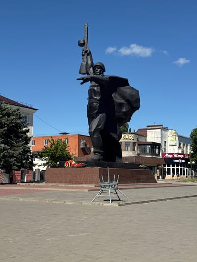 A large, dark bronze statue of a soldier stands in the center of a paved plaza. The soldier is depicted in a dynamic pose, with one arm raised holding a rifle above his head and the other arm extended forward, pointing. He wears a helmet and a cape that billows behind him. At the base of the statue, a wreath of red and yellow flowers rests on a granite pedestal.
Behind the statue, a row of buildings lines the plaza. The closest building on the left is a multi-story structure with a red-tiled roof and light-colored walls. To its right is a brick building with multiple windows, and further to the right are several shops and businesses with awnings and signs. One sign, in Russian, reads "Мир Шмоток" (Mir Shmotok), which translates to "World of Clothes." Another sign has the word "АСС" visible in large gold letters. The sky above is a clear, bright blue with a few wispy white clouds.
In the foreground, to the right of the statue's base, a metal chair is overturned, resting on its side. The plaza pavement is composed of large, grey square tiles. A few small green plants are visible growing in cracks in the pavement. Two people, a man and a woman, are walking away from the viewer on the sidewalk in front of the businesses on the right side of the image.
This statue is the Eternal Flame monument, located in Victory Park in Shakhty, Russia. It commemorates Soviet soldiers who fought in World War II.