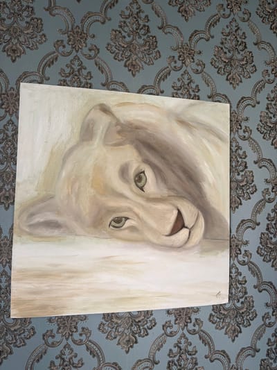 A painting of a lion cub rests on a patterned blue wallpaper. The cub is rendered in light, muted tones of white, beige, and light brown, with green eyes. It appears to be lying on its side, with its head propped up and looking towards the viewer. The background of the painting is a soft, blurred white. The wallpaper features a repeating damask pattern in shades of grey and darker blue. The painting is positioned on the wall, partially covering the wallpaper. A small, faint artist's signature is visible in the bottom right corner of the painting. The scene appears to be an interior domestic setting. There is no specific information to suggest a location in the city of Yuzhny, Russia.