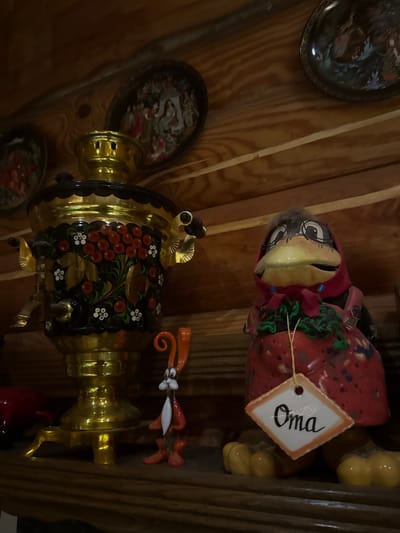 The image depicts a shelf in a room with wooden walls. On the shelf, a brass samovar with floral painted decoration is positioned on the left. To its right, there is a figurine of a bird wearing a red dress and a pink headscarf, with a tag that reads "Oma". A small, orange rabbit figurine stands between the samovar and the bird figurine. Several decorative plates are visible on the wall behind the objects. The scene appears to be indoors, possibly in a home or a decorative shop. The city of Heiligenrode, Germany, is not visually represented or implied in the image.