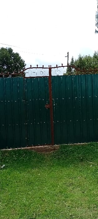 The scene is located in Gatakaine, Kenya. It features a dark green corrugated metal fence with a central, reddish-brown metal gate post. The gate is secured by a dark-colored latch mechanism positioned at its vertical midpoint. The top of the fence is adorned with a light-colored decorative metal railing displaying repeating heart and diamond patterns. White rivets are visible securing the corrugated panels. The foreground consists of dense green grass. In the background, above the fence, are multiple utility wires, dense green foliage from trees, and a wooden utility pole visible on the far right. The sky overhead is overcast. No individuals are present.