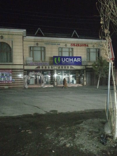 A two-story building at night displays signs for "Wildberries" and "Uchar" (electronics). The building also has "TANTANALAR" and "SHAHZODA" written near the roof. A person walks toward the center entrance under the "Uchar" sign. Ornate decorations frame the Wildberries entrance. The street is wide, dark, and damp. A tree with bare branches stands on the right side of the frame. Wires stretch across the dark sky.