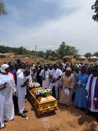 A large group of people is gathered outdoors, likely in Dogon Dinia, Nigeria, near a wood coffin decorated with flowers. Many individuals are dressed in white, while others wear a variety of colors and patterns. A priest stands near the coffin wearing a purple and white robe with a cross symbol. The environment includes dirt ground, trees, houses, and power lines, suggesting a rural setting.
