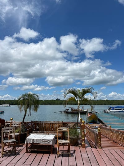 The image depicts a waterfront scene in Ko Panyi, Thailand, on a bright, partly cloudy day. The foreground is a wooden deck furnished with rustic wooden chairs and a table covered with a white, patterned cloth. Two palm trees are visible on the deck, adding a tropical feel to the setting. Beyond the deck, a body of calm water stretches out, with several boats moored along a blue pier. The far bank of the water is lined with a dense green forest, and a faint outline of mountains can be seen in the distance under the expansive blue sky dotted with white cumulus clouds. The overall atmosphere is serene and inviting, suggesting a peaceful day by the sea.
