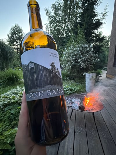 A person's hand holds a partially filled bottle of "LONG BARN ZINFANDEL CALIFORNIA" wine outdoors on a wooden deck. In the background, a metal fire pit contains a burning fire with visible flames and embers. Green foliage, trees, and a white corrugated trash bin are also present in the environment. A portion of a brick building is visible to the right. The sky above is clear and blue. The location is not specified in the image or crops provided.