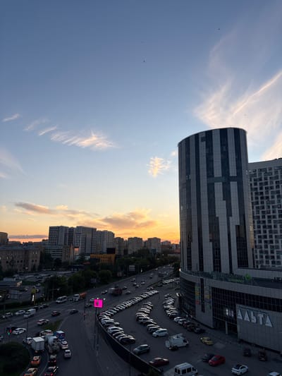 This is a high-angle, landscape-oriented photograph taken from a rooftop overlooking a cityscape at dusk. The sky is a gradient of deep blue transitioning to a warm orange and yellow glow near the horizon, indicative of sunset. Wispy clouds catch the fading sunlight, creating streaks of light and shadow.

The foreground is dominated by a large, modern building with a curved glass facade, reflecting the sky and surrounding architecture. To its left, the sky takes up a significant portion of the frame, with a few small birds visible in flight. To the right, the building's facade features a grid pattern of dark and light panels. A large sign with white lettering, reading "АШАН" (ASAN) is visible on the building's exterior.

Below the buildings, a busy street is visible, filled with numerous cars parked along the roadside and moving in traffic. The vehicles are varied in color and type, including cars, trucks, and vans. The street is illuminated by streetlights and the ambient glow of the sunset. Further into the distance, a dense arrangement of apartment buildings and other structures forms the urban landscape. Trees and green spaces are interspersed among the buildings, particularly along the roadside.

The time of day is clearly sunset, evidenced by the dramatic sky colors. The weather appears clear with some cloud cover. The presence of the "ASAN" sign and the architecture suggest an urban or suburban commercial area in Russia, consistent with the provided location context of Lesnyye Polyany. The overall mood is serene, with the beauty of the sunset juxtaposed against the activity of the city.