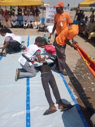 The scene takes place in Katabu, Nigeria at the coordinates 10.71 latitude and 7.514 longitude. A man wearing a red shirt with a white cross is kneeling next to an injured man lying on a white tarp. The injured man has dark skin and is wearing black pants. Two other men wearing orange shirts and pants are standing to the right of the scene. One of the men is holding a stretcher. A woman wearing a white shirt with a red cross is kneeling in the background. In the distance, there are several people sitting on chairs under a yellow tent. The scene is brightly lit and there is a blue and white tarp in the foreground. The man in the red shirt with the cross is tending to the injured man while the two men in orange shirts are observing the scene. The woman is watching the scene unfold in the distance.