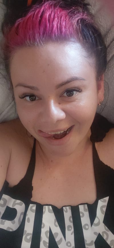 A woman with pink and black hair, wearing a black top with "PINK" written on it in a leopard print font, is lying down and looking at the camera. Her tongue is sticking out and touching her lips. The image is a selfie, taken from a close-up perspective. The background appears to be a bed or couch. There are no discernible landmarks or contextual clues to indicate the location as Marynivka, Ukraine.