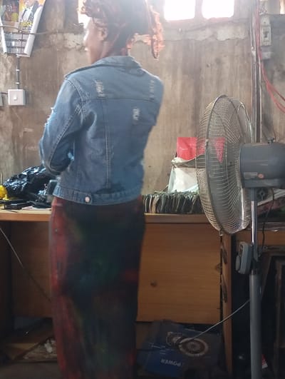 A medium shot captures a woman from behind, standing in what appears to be a cluttered workshop or market stall. She wears a light blue denim jacket with distressed detailing and a long, colorful skirt with abstract patterns of red, green, and black. Her hair is tied up, and she wears a patterned headscarf.
The setting is dimly lit, with natural light filtering in from a window above and to the right, creating a bright glare that obscures details in that area. The walls are rough and textured, suggesting an unfinished or rustic interior. In the background, a large, oscillating metal fan stands on a stand. To the left of the woman, a desk or counter is visible, laden with items including a black plastic bag, what looks like a power strip, and a stack of papers or books. A poster with text is partially visible on the wall above the desk. A power cord snakes across the floor and up a wall.
The woman's posture is relaxed as she faces away from the camera. The overall impression is one of everyday activity within a working environment. The time of day is indeterminate due to the lighting. There are no overt cues about the weather or a specific event. The visible text on the poster is too small and out of focus to be legible. The scene is likely indoors, given the presence of the fan and the construction of the walls.