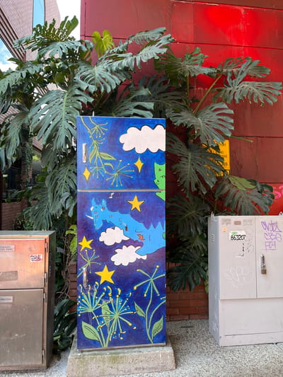 This vertical media file captures a vibrant urban scene in South Brisbane, Australia, dominated by lush foliage and colorful street art on utility infrastructure.

The primary subject is a large, vigorous Monstera deliciosa or similar tropical plant, with numerous large, fenestrated green leaves, cascading and climbing against a deep red, paneled wall. The plant covers a significant portion of the wall, with some leaves extending over the painted utility cabinet.

In the center of the frame is a rectangular utility cabinet, divided into two panels, which has been artfully painted. The artwork depicts a stylized night sky against a dark blue background. White, puffy clouds and bright yellow four-pointed stars are scattered across both panels. Additionally, delicate green leafy patterns, resembling dill or similar flowering plants, adorn the dark blue sky. The right panel also features abstract light blue shapes, vaguely resembling landmasses or an island group, further contributing to the sky-and-land motif. Each panel has a small, silver handle.

Above the painted cabinet, attached to the red wall, is a grey metal utility box covered in graffiti and a white sticker displaying the number "863207". Below the painted cabinet, another grey metal utility box is visible, also with some text and labels such as "Go... HASH", "ENERGY METER PANEL", and partially visible "TRIBUTION". A small yellow sign on the red wall behind the plant is partially obscured, showing text like "RTPORT", "LANS... BRIDGE", and "303", possibly indicating a location or postcode.

The scene is brightly lit, suggesting daytime, contrasting with the night-themed painting. The combination of dense natural growth, functional urban infrastructure, and artistic expression creates a lively and visually engaging streetscape.