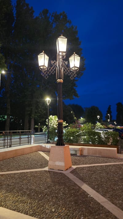 The image shows an outdoor scene at dusk or night, likely in Peschiera del Garda, Italy, given the context. The primary subject is a decorative streetlamp with multiple illuminated glass fixtures, casting a warm glow. The lamp post is ornate and made of dark metal, featuring geometric decorative elements.

The setting appears to be a pathway or plaza paved with small, dark pebbles interspersed with light-colored lines, creating a patterned effect. To the left of the streetlamp, a large, dark green tree with dense foliage is visible against the deep blue twilight sky. Beyond the tree and lamp post, there are glimpses of other streetlamps, greenery, and what appears to be a building or wall.

In the background, further illuminated trees and flowering plants suggest a park or garden area. A low, decorative metal railing can be seen along the edge of the pathway. The time of day is clearly indicated by the dark sky and the artificial lighting, suggesting it is after sunset. The weather appears calm and clear.

No people are visible in the image, and the focus is on the architectural and natural elements of the scene. The overall mood is serene and peaceful, enhanced by the gentle lighting and the stillness of the evening. There is no visible text in the image.