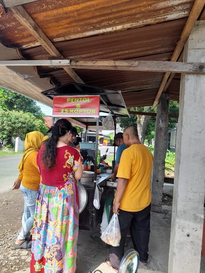 Captured in Waung, Indonesia, the image depicts a street scene centered around a food stall. The stall, labeled "PENTOL & ES KOPYOR," is the focal point. Two individuals are interacting with the stall; one, wearing a yellow shirt and black pants, is holding a plastic bag. Another person, dressed in a colorful batik-patterned top, is positioned closer to the stall. A third person wearing yellow is standing behind the batik-patterned top person. The surrounding environment features a mix of corrugated metal roofing, concrete structures, and street vegetation.