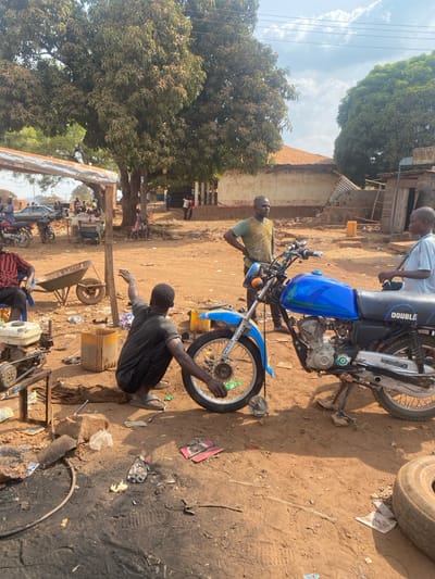 An outdoor scene in Nev, Nigeria, depicts an informal motorcycle repair area under bright daylight. In the foreground, a dark-skinned male wearing a dark t-shirt and dark trousers is squatting, actively engaged in repairing the front wheel of a blue and black motorcycle, model "DOUBLE HORSE." His hands are positioned on the wheel assembly, which is elevated. A silver brake drum component lies on the dusty ground directly beside the wheel.

To the right of the motorcycle, a dark-skinned male in a light green/khaki t-shirt with "ORIS" branding stands with his hands on his hips, observing the repair. Further to the right, a younger dark-skinned male in a light blue t-shirt is partially visible, also standing near the motorcycle.

On the left side of the frame, a dark-skinned male in a red and black striped shirt is seated on a red plastic chair beneath a rudimentary canopy structure, composed of metal poles and a light-colored sheet. Behind him, a metal wheelbarrow is visible. A small, generator-like engine sits on a wooden stand in the immediate left foreground.

The ground consists of reddish-brown dusty soil, with scattered debris including metal parts, plastic bags, and a discarded tire. A dark, oily stain is present on the ground in the foreground. In the mid-ground, a yellow jerrycan and a wooden crate are visible.

The background features several large trees with dense green foliage. To the left, additional motorcycles and vehicles are parked, with more individuals visible in the distance. To the right, a light-colored building with a terracotta-colored corrugated metal roof stands, alongside a partially collapsed or dilapidated structure. Overhead power lines are visible against a sky with scattered white clouds. The overall environment suggests an active, bustling outdoor workshop or market setting.