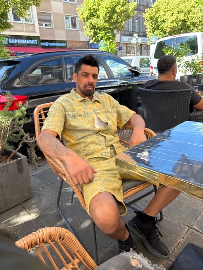 A man with a goatee and dark, spiky hair sits in a wicker chair at an outdoor cafe, facing the camera. He is wearing a yellow and white tie-dye short-sleeved shirt and matching shorts. His left arm, which rests on his lap, displays a tattoo. To his right, a dark table with a reflective surface is visible. In the background, a black car is parked, and beyond that, buildings with signs for "SUPERMARKT" are visible. Trees with green foliage frame the scene. Another person, wearing a black shirt, sits in a dark chair at a table further back. The ground is paved with gray square tiles. The image appears to be taken on a sunny day in Nuremberg, Germany.