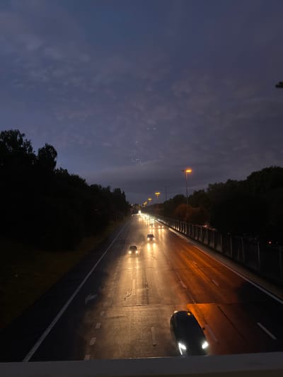 The image depicts a multi-lane highway in Anderlecht, Belgium, viewed from an elevated vantage point during twilight or night conditions. The sky above is dark blue-grey with visible cloud formations, featuring a faint, dispersed cluster of small, bright points in the upper mid-right section. The road surface is wet, highly reflective, and illuminated by both vehicle headlights and orange-yellow streetlights. Multiple vehicles are in motion, traveling away from the viewer in both visible lanes, their headlights casting bright, elongated reflections on the damp asphalt. A dark vehicle with prominent white LED headlights is partially visible in the immediate foreground, positioned on the far right lane. The left side of the highway is bordered by a dark, densely wooded embankment. The right side features a concrete or metallic barrier running parallel to the road, with additional dark foliage visible behind it. No individuals are present in the scene.