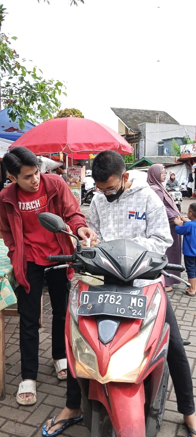 The image depicts two young men standing by a red Honda motorcycle parked in front of a small shop in Banjaran, Indonesia, located at latitude: -7.046, longitude: 107.581. The man on the left is wearing a red jacket, a red T-shirt, black jeans, and white sandals. The man on the right is wearing a white and grey patterned jacket with a black mask, black pants, and blue flip-flops. The motorcycle has a red license plate that reads "D 6762 MG 10.24". A woman in a hijab is standing behind the motorcycle, and a young boy in a blue shirt is standing to the right of the motorcycle, looking at the camera. There is a red umbrella in the background, partially obstructing the view of a small shop. The sky is overcast. The image appears to be taken on a street with brick pavement, where the men are looking at the motorcycle.