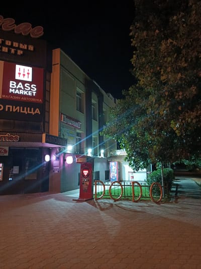 The image depicts a street scene at night in Volzhsky, Russia, specifically on Prospekt Lenina. The main subjects are commercial establishments and their signage, illuminated by artificial lights.
On the left, a building features prominent signs. The topmost sign, partially visible, appears to be a neon sign with large, stylized letters that are difficult to decipher fully but suggest a name like "Tsar." Below this, another sign reads "ТОГОВЫЙ ЦЕНТР" (Shopping Center). Further down, a bright red sign with white lettering declares "BASS MARKET" with the tagline "МАГАЗИН АВТОЗВУКА" (Car audio store). Adjacent to this, a sign with large orange letters says "О ПИЦЦА" (About Pizza). Below these, a dark facade with illuminated windows suggests another storefront, with a partial sign visible that reads "ovies." Further to the left, a black sign with white text is partially visible.
In the center of the image, a modern red vending or service kiosk with a digital screen stands on the paved walkway. It has branding that includes "RICKY BOXER" and Cyrillic text. Behind this kiosk, a metal fence encloses a grassy area with some bushes.
To the right of the kiosk, a building with multiple windows is visible, its facade featuring horizontal grey paneling. Signs on this building include one that reads "Приоритет" (Priority) and another with "ZEBRA-TOUR".
A large tree with green and yellowing leaves dominates the right side of the frame, casting shadows on the ground. The walkway in the foreground is paved with brick-like tiles. Bicycle racks, designed as orange circles, are positioned along the fence. The overall impression is that of a commercial street at night, with businesses open or lit, and a moderate amount of pedestrian traffic implied by the presence of shops and walkways. No people are clearly visible in the image.