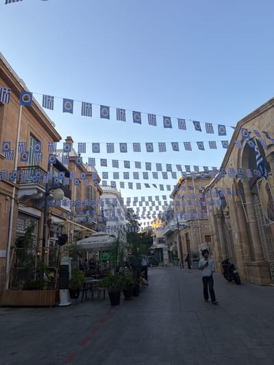 The image depicts a street scene decorated with numerous small Greek flags strung across the sky, creating a festive atmosphere. The flags are predominantly white and blue, with a smaller blue flag featuring a white cross and alternating blue and white stripes, and a separate blue flag with a circular emblem. Buildings with traditional architecture line the street, some featuring balconies and ornate details.

Several people are visible in the scene. In the foreground, a man wearing a turban and casual clothing stands on the cobblestone pavement, looking at his phone. Further down the street, a few individuals are seated at outdoor tables, suggesting a cafe or restaurant setting. Two people are walking on the sidewalk, one in the distance and another closer to the flags.

The time of day appears to be late afternoon or early evening, as indicated by the warm, soft light and the lengthening shadows on the pavement. The sky is clear and blue, suggesting fair weather. The presence of Greek flags strongly hints at a celebration or a significant national event in Cyprus, possibly related to Greek heritage or independence. The overall mood conveyed is one of pleasantness and community engagement. The scene is bustling with activity, yet also has a relaxed ambiance, typical of a European street in the evening.