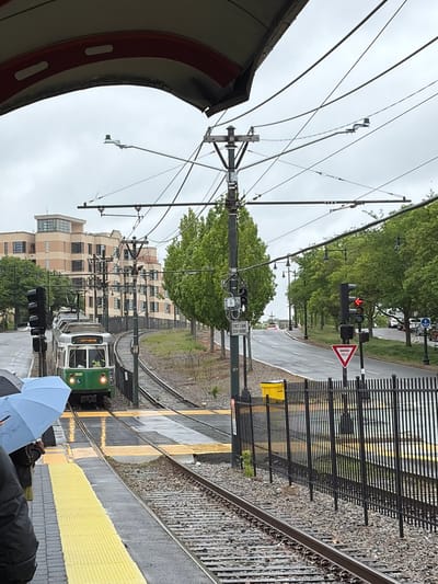 The image depicts a rainy day scene at a platform of the MBTA Green Line, likely the C branch, in Boston, Massachusetts. A green and silver trolley is pulling into a station, with its destination sign reading "GOVT CENTER." Overhead wires supply power to the train. A light blue umbrella is visible, suggesting people are waiting on the platform. A street runs parallel to the train tracks, equipped with traffic lights and yield signs, and lined with trees and adjacent buildings. The overall atmosphere is overcast and wet.