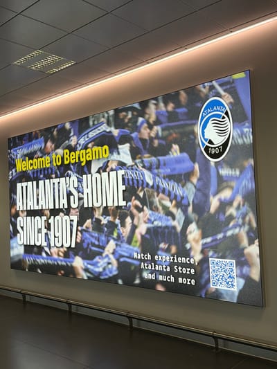The image shows a large digital billboard advertisement for Atalanta, a football club, located in the city of Orio al Serio, Italy, likely at the airport. The billboard features a crowd of fans wearing blue and black scarves, with the Atalanta logo prominently displayed in the upper right corner. Text on the billboard reads "Welcome to Bergamo," "ATALANTA'S HOME," and "SINCE 1907." Below this, smaller text says "Match experience, Atalanta Store and much more," alongside a QR code. The billboard is mounted on a wall with a metallic railing in front of it, and a tiled floor below. The environment suggests an indoor public space, such as a terminal.