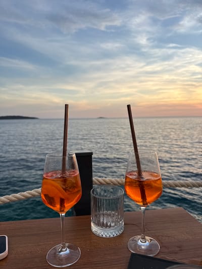Two glasses of orange aperitif drinks with straws sit on a wooden table overlooking the sea at sunset in Primošten, Croatia. The sky is a blend of blue, pink, and orange hues, with wispy clouds. The sea is calm, with gentle waves. In the foreground, a thick rope runs horizontally along the edge of the table. A clear glass with a ribbed texture is positioned between the two drinks. A dark object, possibly a wallet or phone case, rests on the table in the lower right corner.