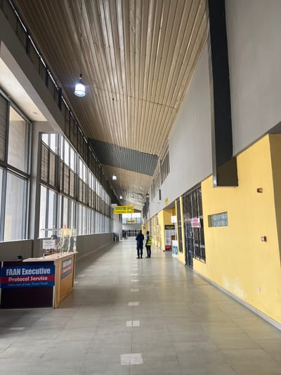 The image depicts an interior space, possibly an airport terminal, located in Kwi, Nigeria. Two individuals wearing what appears to be security vests are positioned further down the hallway. On the right side of the hallway, there is a booth labeled "FAAN Executive Protocol Service." A sign above indicates "DEPARTURE HALL SCREENING POINT." The architecture is marked by a high ceiling, metal siding on one side, and a window-lined wall with multiple panes on the other. The floor is light-colored and appears to be made of tile or a similar material.