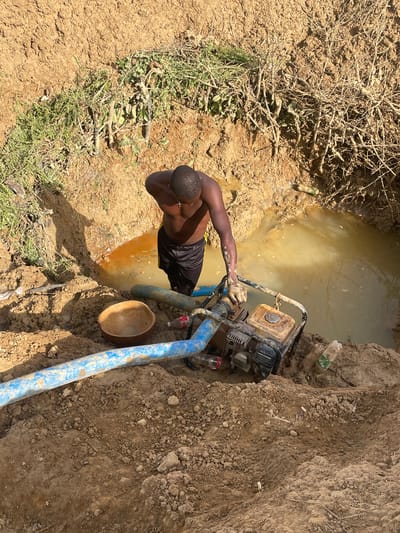 In an outdoor environment near Gana Daji, Nigeria, an individual with dark skin, wearing black shorts and no shirt, stands beside a muddy hole filled with murky water. The person appears to be operating a gasoline-powered water pump, indicated by the hoses and the visible engine. The pump sits close to the edge of the hole. A wooden bowl rests on the ground next to the blue pump hose. The immediate surroundings include soil, sparse vegetation, twigs, and discarded plastic bottles. The landscape appears dry and dusty.
