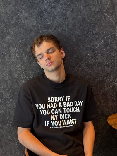 An adult male is depicted resting against a dark, textured wall in Minhang District, China. He has brown, wavy hair and his eyes are closed with his lips slightly parted, suggesting a state of sleep or deep relaxation. He is wearing a black, short-sleeved crew-neck t-shirt with white text printed prominently across the chest. The text reads: "SORRY IF / YOU HAD A BAD DAY / YOU CAN TOUCH / MY DICK / IF YOU WANT". Below this, a website address "www.mimimamam.com" is visible. The wall behind him features a dark gray, stone-like surface with subtle lighter veining. A small portion of a polished wooden surface, consistent with a table or chair arm, is visible in the lower right corner of the frame. The lighting is even, illuminating the man's face and shirt without harsh shadows.