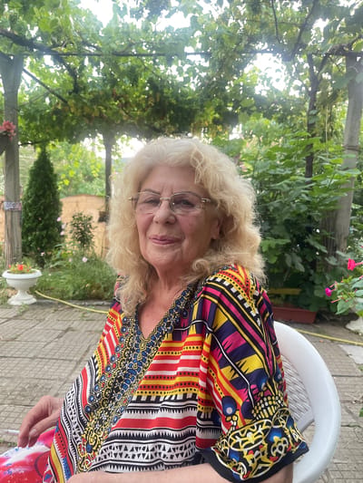 The image shows a woman with blonde hair and glasses, wearing a colorful, patterned shirt. She is seated in a white plastic chair outdoors, with a lush green garden visible behind her. The sun appears to be shining, casting dappled light through the leaves of trees and vines. The ground is paved with light-colored tiles. In the background, there are more trees, bushes, and some pink flowers in a white planter. A yellow hose is also visible on the ground. The overall impression is that of a casual outdoor setting, likely a garden or patio. The woman's expression appears relaxed. The time of day is likely daytime, given the bright lighting. There is no visible text in the image. The location is Simitli, Bulgaria.