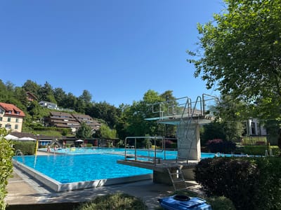 The photograph captures an outdoor swimming pool facility on a clear, sunny day. In the foreground, a bright blue swimming pool dominates the scene, with a diving board structure to the right. The diving board has multiple levels and metal railings, with a ladder visible on its side. The pool area is surrounded by concrete pathways and green foliage, including bushes and trees. In the background, houses are visible on a hillside, with a clear blue sky above. Several people are present in the pool, some swimming and others relaxing on inflatables. There are also some umbrellas providing shade. The overall environment appears to be a recreational public space. The provided coordinates indicate that this location is in the city of Wald (ZH), Switzerland.
