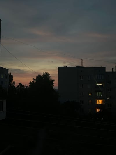 The image depicts a twilight scene in Peremyshliany, Ukraine, characterized by the interplay of fading daylight and artificial illumination. In the foreground, several dark, horizontal wires stretch across the frame, silhouetted against the dusky sky. These wires are likely part of the city's infrastructure.
Dominating the mid-ground are the dark silhouettes of trees, their dense foliage obscuring whatever lies behind them. To the left, the partial facade of a grey, multi-story apartment building is visible, its windows dark except for a faint, green light emanating from one.
Further to the right, a larger, darker apartment building stands prominently. Several windows in this building are illuminated with a warm, orange glow, suggesting interior lights are on. These lights cast distinct beams and reflections, particularly from the lower floors. The building is topped with what appear to be television antennas. The sky above is a gradient of muted greys and purples, with subtle hints of pink and orange near the horizon, indicative of a sunset or post-sunset sky. The overall atmosphere is one of quiet dusk in a residential urban environment.