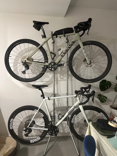 The image shows two bicycles mounted on a wall rack. The upper bicycle is a gravel bike with a light green frame and black handlebars. It has a saddle bag and a water bottle cage. The lower bicycle is a road bike with a white frame and black handlebars. It also has a water bottle cage and the brand name "BELLE" is visible on the frame. Both bicycles are equipped with disc brakes. The background appears to be a wall in an indoor setting, possibly a garage or storage area. A Monstera plant is visible to the right. The image does not provide any information about the location being Vilanova i la Geltrú, Spain.