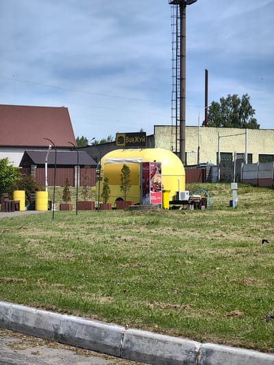 A bright yellow, rounded food truck, branded with a burger logo and the text "BURЖУИ", is parked on a grassy area in front of a light yellow brick building. The food truck has a Coca-Cola advertisement on its side. To the left of the food truck, there are two bright yellow barrels and some wooden crates. In the foreground, there is a gray concrete curb and a grassy lawn. In the background, a tall metal structure with a ladder and a reddish-brown smokestack are visible against a cloudy blue sky. The scene appears to be in a semi-urban or industrial area.