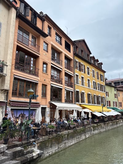 This is a vertically oriented, eye-level, full shot of a canal-side building facade in Annecy, France. The buildings are tall and narrow, with multiple stories featuring numerous windows. The facade colors range from a warm, earthy orange on the lower floors to a bright yellow and a pale green on the upper levels. The architecture is old, with wooden shutters and iron grates visible on many windows.

Along the canal, an outdoor seating area for a restaurant is set up. Several people are seated at tables, enjoying meals and drinks. White umbrellas and awnings provide shade. The canal itself is visible on the right side of the image, with textured stone retaining walls. The water appears murky green.

The sky is overcast, suggesting a cloudy day. The time of day appears to be daytime, possibly late morning or afternoon. A street lamp is visible in the foreground. Some plants and greenery are placed around the outdoor seating area, adding a touch of life to the scene. Text visible on an awning reads "BRUNCH ALL DAY". The overall atmosphere seems relaxed and leisurely, typical of a European canal-side setting.
