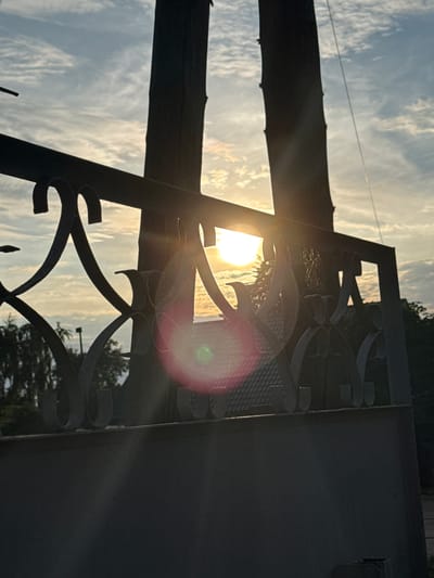 The image shows a sunset behind a fence with ornate scrollwork. The sun is bright and causes lens flare, creating a pink and green circular glow. Two dark, vertical structures, possibly fence posts or poles, are silhouetted against the sky. The sky is partly cloudy with streaks of white and grey clouds. In the background, trees and a building roof are visible. The foreground consists of a dark, solid fence or wall. There are no people visible in the image. The scene appears to be outdoors, likely in a residential or park area. The location is Rzhev, Russia, as indicated by the request.