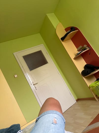 An interior shot shows a room from an unusual, rotated perspective, with a person's arm and leg (wearing light blue ripped denim shorts) partially visible on the right side. The dominant wall color is lime green. A white door with a frosted glass window and a handle is visible on what appears to be a wall, though its orientation in the image makes it seem horizontal. Above or beside the door, a multi-colored shelf unit is mounted, featuring a reddish-brown backboard and light yellow shelves with rounded edges. Items on the shelves include a pair of black boxing gloves, two pairs of black shoes or slippers, a bright green plastic basket, and some electronic devices or cables. The floor, partially visible under the person's leg and to the right, consists of a light yellow section, wooden laminate flooring, and a striped orange and brown rug. The scene depicts an interior space, likely a home, with a vibrant color scheme. There is no visible text.