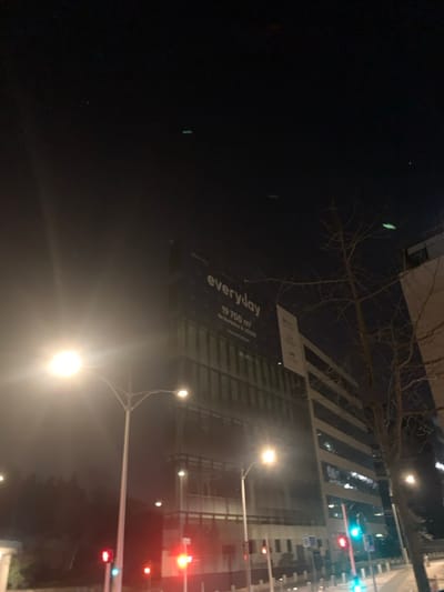 This night-time image depicts an urban commercial area within or immediately bordering Issy-les-Moulineaux, France. The central subject is a multi-story modern office building, partially obscured by a prominent dark banner across its upper facade. The banner displays the word "everyday" in white lettering, followed by "19 700 m²" and the French phrase "de bureaux à LOUER" (office space for rent). Below this text are several smaller, indistinct logos. To the right of this building, another structure with horizontal bands and visible interior lighting is present.

The scene is primarily illuminated by artificial light sources. Multiple bright streetlights are visible in the foreground and midground, generating significant lens flare. Traffic lights are visible at street level; those on the left display red, while a signal further right shows a green light. Leafless deciduous trees are visible on the right side of the frame, and darker foliage lines the far left. Low barriers or bollards are present along what appears to be a curb or median in the foreground. No individuals, specific actions, or interactions are discernible.